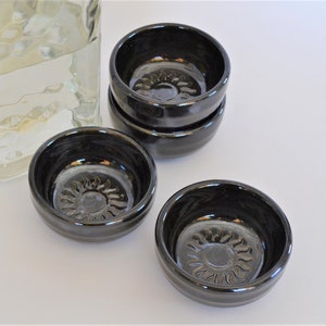 May include: Four small black ceramic shot glasses with a sun design on the bottom. The glasses are stacked on top of each other.
