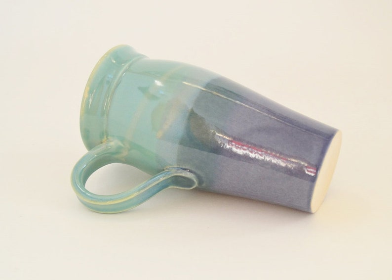 May include: A ceramic mug with a light blue to dark blue ombre glaze. The mug has a handle and a rounded shape.