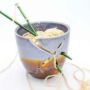 May include: A ceramic yarn bowl with a blue and brown glaze. The bowl has holes for the yarn to feed through. A ball of white yarn is in the bowl, and two green knitting needles are sticking out of the yarn.