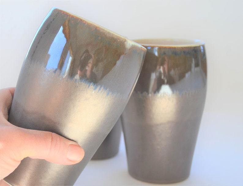 May include: Two ceramic mugs with a brown and white glaze. The mugs have a rounded shape and a wide base.