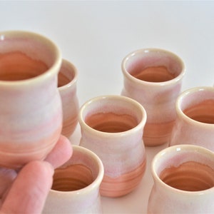 May include: Set of eight small, hand-thrown ceramic cups in a light pink and cream glaze. The cups have a rounded shape and a slightly flared rim.