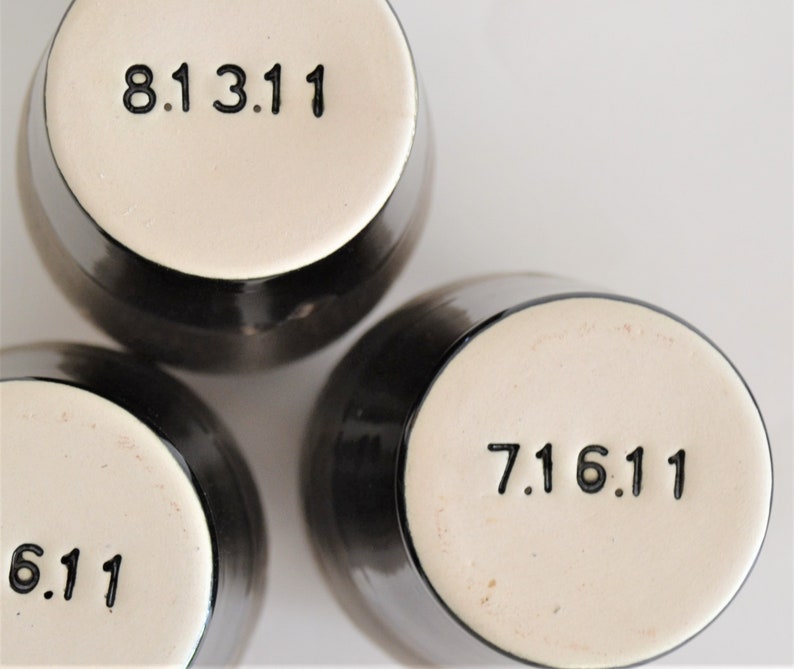 May include: Three black ceramic mugs with white bottoms. Each mug has a black number printed on the bottom. The numbers are 8.13.11, 7.16.11, and 6.11.