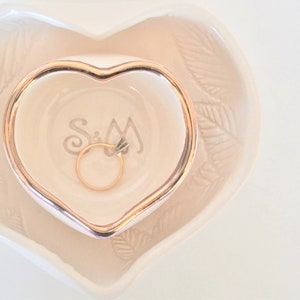 May include: A white ceramic heart-shaped dish with a gold rim. The dish has a smaller heart-shaped dish inside with a gold rim. The smaller dish has the initials "S&M" inscribed on the bottom and a gold ring inside.