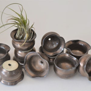 May include: A collection of ten small, brown ceramic pots with a textured, metallic finish. Some of the pots have a drainage hole. One pot is taller and has a plant growing in it.