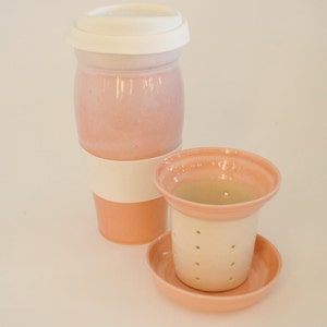 May include: A pink ceramic travel mug with a white lid and a white band around the middle.  The mug is next to a pink ceramic tea infuser with a white strainer and a matching saucer.