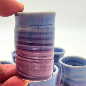May include: A handmade ceramic shot glass with a gradient of colors, transitioning from blue to purple and pink. The shot glass has a cylindrical shape and a textured surface. Several similar shot glasses are visible in the background.