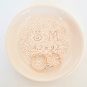 May include: A white ceramic ring dish with a rose pattern and the initials "S & M" and the date "4.26.92" engraved on the surface. Two gold rings are resting on the dish.