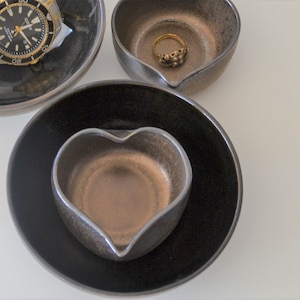 May include: Three heart-shaped ceramic dishes, one black, one brown, and one with a black rim and a brown interior. The brown dish is nested inside the black dish. A gold ring is in the top brown dish.