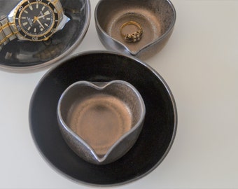 Puede incluir: Tres platos de cerámica en forma de corazón, uno negro, uno marrón y uno con borde negro e interior marrón. El plato marrón está anidado dentro del plato negro. Un anillo de oro está en el plato marrón superior.