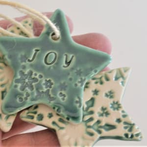 May include: Three ceramic star ornaments with a green glaze and white snowflake pattern. One star has the word "JOY" written on it.