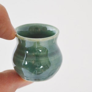 May include: A small, green ceramic pot with a rounded body and a slightly flared rim. The pot is glazed in a light green color with a subtle sheen.