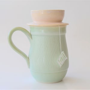 May include: A light green ceramic tea pitcher with a lid and a tea bag hanging from the handle. The pitcher has a textured, wavy design.