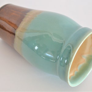 May include: A ceramic vase with a glossy finish. The top section is brown, transitioning to tan, then a teal-green body. The interior has a light orange hue. The vase has a wide opening and a curved shape.