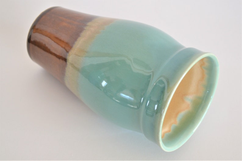 May include: A ceramic vase with a glossy finish. The top section is brown, transitioning to tan, then a teal-green body. The interior has a light orange hue. The vase has a wide opening and a curved shape.