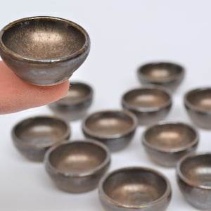 May include: A set of ten small, round, brown ceramic bowls with a metallic sheen. The bowls are arranged in a semi-circle on a white surface.