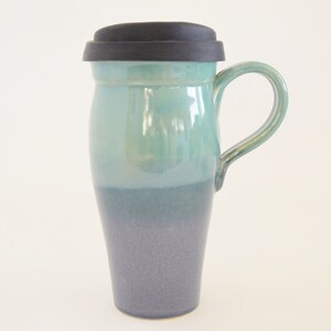 May include: A ceramic travel mug with a teal and dark blue ombre glaze. The mug has a black silicone lid and a handle.