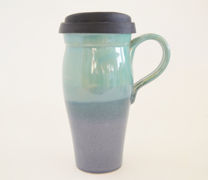 May include: A ceramic travel mug with a teal and dark blue ombre glaze. The mug has a black silicone lid and a handle.