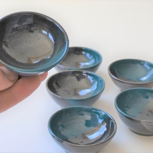 May include: Six small, teal and black ceramic bowls. The bowls are round and have a glossy finish. They are arranged in a row on a white surface.