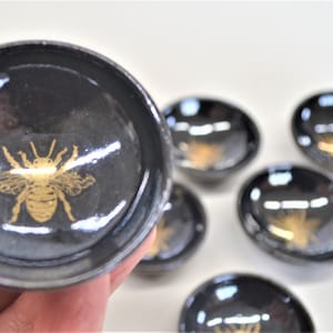 May include: Set of seven small black ceramic dishes with gold bee designs. The dishes are perfect for serving small snacks or condiments.