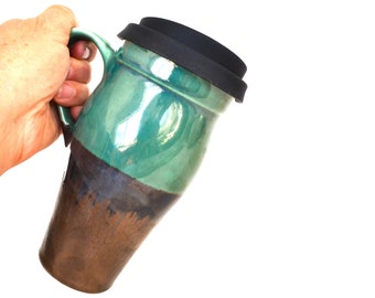 May include: A green and brown ceramic travel mug with a black lid. The mug has a unique glaze pattern and is held in a hand.