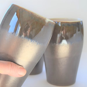 May include: Two ceramic mugs with a brown and white glaze. The mugs have a rounded shape and a wide base.