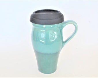 May include: A teal ceramic travel mug with a black silicone lid. The mug has a rounded body and a handle.