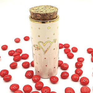May include: A cream-colored ceramic jar with a gold heart and arrow design, topped with a cork stopper. The jar is surrounded by red candies. The jar has gold polka dots and a gold rim.