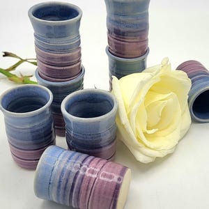 Peut inclure: Ensemble de petits verres à liqueur en céramique faits à la main, dans des tons de bleu et de violet. Les tasses cylindriques ont un design texturé et nervuré. Une rose jaune est disposée parmi les verres.
