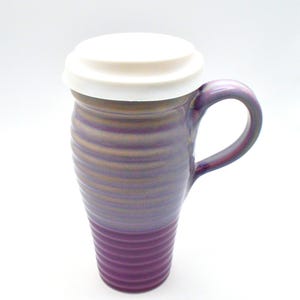 May include: A tall, purple ceramic travel mug with a white lid. The mug has a handle and a ridged texture. The color gradient transitions from a deep purple at the base to a lighter purple and gold near the top.