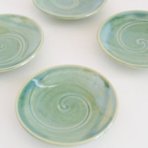 May include: Four small, green ceramic dishes with a swirling pattern. The dishes are on a white surface.