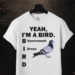 以下が含まれることがあります： 中央にYEAH, I'M A BIRD. B Government I Drone R Dという黒い文字と、横向きに立つ灰色で虹色に輝く鳩のリアルなカラー写真がプリントされた、白い半袖のTシャツ。