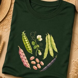 Può includere: Una t-shirt verde bosco scuro con un'illustrazione botanica di vari legumi. La grafica include un baccello di piselli, un baccello di fagioli viola, un baccello scuro, diversi fagioli maculati sparsi e un viticcio con piccoli fiori bianchi.