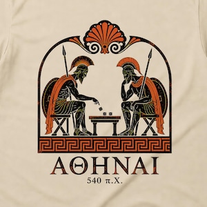May include: A beige short-sleeve t-shirt featuring a black and orange graphic of two ancient Greek warriors playing a game at a table. The design includes a decorative arch, a Greek key border, and the text "AOHNAI 540 π.X." printed below the illustration.