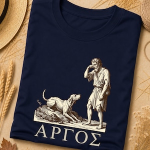Puede incluir: Una camiseta azul marino con una ilustración gráfica de color crema de un hombre de pie junto a un perro. El hombre se toca la cara y el perro está tumbado. Debajo de la ilustración, el texto griego "ΑΡΓΟΣ" está impreso en letras blancas.