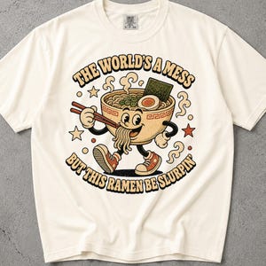 May include: An off-white t-shirt featuring a cartoon ramen bowl with arms and legs wearing trainers, holding chopsticks. The text reads: THE WORLD'S A MESS BUT THIS RAMEN BE SLURPIN'. The graphic includes steam, stars, and small circles.