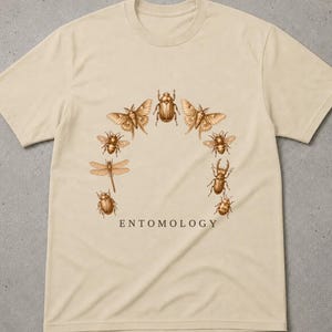 Könnte beinhalten: Ein beiges Kurzarm-T-Shirt mit einer kreisförmigen Anordnung verschiedener brauner Insektenillustrationen, darunter Käfer, Motten und eine Libelle. Das Wort ENTOMOLOGY ist in einer dunkelbraunen Serifenschrift unter den Insekten gedruckt.