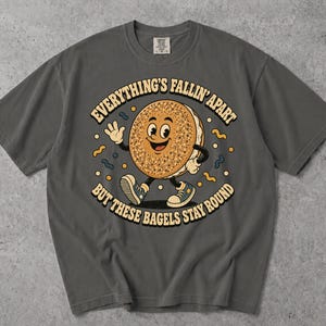 May include: A charcoal grey t-shirt featuring a cartoon bagel character with arms and legs wearing trainers. The graphic includes the text "EVERYTHING'S FALLIN' APART BUT THESE BAGELS STAY ROUND" surrounded by small yellow and blue abstract shapes.