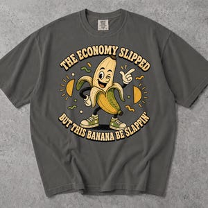 May include: A charcoal grey short-sleeve t-shirt featuring a cartoon banana character wearing green trainers. The graphic includes the text THE ECONOMY SLIPPED BUT THIS BANANA BE SLAPPIN, surrounded by yellow sun icons and small green and yellow squiggles.