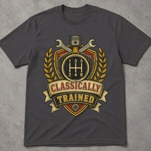 May include: A charcoal grey t-shirt featuring a graphic of a manual transmission gear shift pattern, a piston, and two crossed wrenches inside a shield emblem. Below the emblem, a banner displays the text "CLASSICALLY TRAINED" in bold letters.
