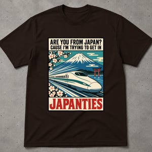 May include: A dark brown short-sleeve t-shirt featuring a graphic print of a high-speed train, Mount Fuji, cherry blossoms, and a red torii gate. The text reads: ARE YOU FROM JAPAN? CAUSE I'M TRYING TO GET IN JAPANTIES in black and bold red lettering.