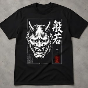 Könnte beinhalten: Ein schwarzes Kurzarm-T-Shirt mit einem weißen Grafikdruck einer Hannya-Maske mit Hörnern. Das Design enthält japanische Kanji-Schriftzeichen, einen kleinen japanischen Textblock und einen roten quadratischen Stempel mit weißen Schriftzeichen.