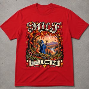 Può includere: Una t-shirt rosso brillante con un'illustrazione grafica di un mago con una tunica blu e una volpe in un bosco autunnale con foglie d'acero, una cascata e montagne. Il testo "MILF" appare in alto e "Man I Love Fall" nella parte inferiore.