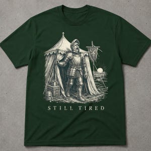 Könnte beinhalten: Ein dunkelgrünes T-Shirt mit einer detaillierten weißen Grafik eines Ritters in vollständiger Plattenrüstung, der am Eingang eines Zeltes steht. Der Ritter scheint zu gähnen. Unter der Abbildung steht der Text STILL TIRED in Serifenschrift.