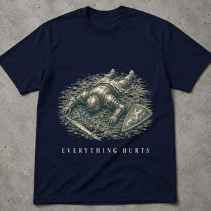 May include: A navy blue short-sleeve t-shirt featuring a graphic illustration of a fallen knight in armour lying on the ground with a sword and shield. The text "EVERYTHING HURTS" is printed in white capital letters below the central graphic.