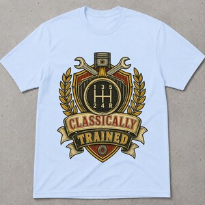 May include: A light blue t-shirt featuring a graphic print of a shield with a manual transmission gear shift pattern, a piston, and two crossed wrenches. The design includes golden laurel leaves and a banner with the text CLASSICALLY TRAINED.