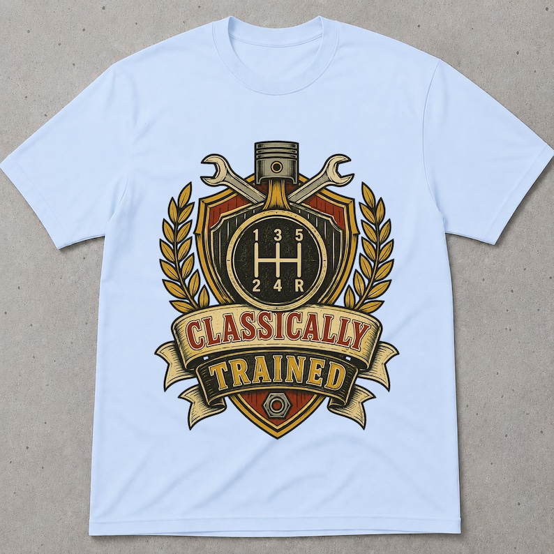 May include: A light blue t-shirt featuring a graphic print of a shield with a manual transmission gear shift pattern, a piston, and two crossed wrenches. The design includes golden laurel leaves and a banner with the text CLASSICALLY TRAINED.