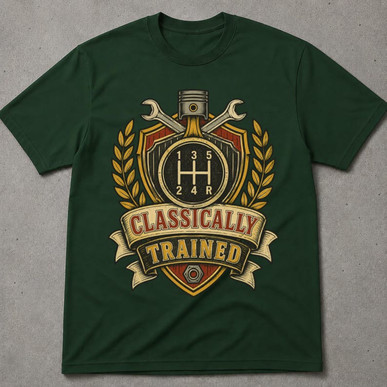 May include: A dark green short-sleeve t-shirt featuring a graphic print of a car gear shift pattern, a piston, and two wrenches arranged on a shield. The design includes yellow wheat stalks and a banner with the text "CLASSICALLY TRAINED".