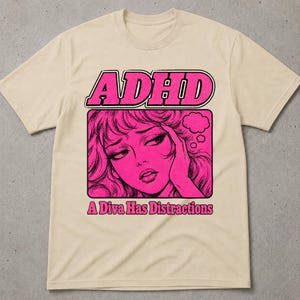 以下が含まれることがあります： 鮮やかなピンク色のグラフィックプリントが施されたベージュの半袖Tシャツ。上部に「ADHD」の文字、頭に手を当てた人物のスタイライズされたイラスト、下部に「A Diva Has Distractions」という文字がデザインされています。