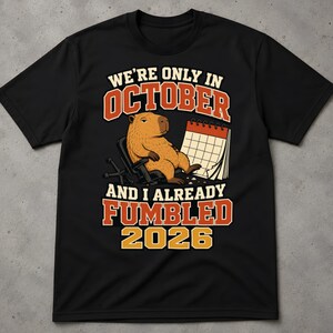 Può includere: Una t-shirt nera a maniche corte con la grafica di un capibara marrone seduto su una sedia accanto a un calendario. Il testo recita: WE'RE ONLY IN OCTOBER AND I ALREADY FUMBLED 2026. Il design utilizza scritte di colore arancione, bianco e giallo.