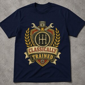 May include: A navy blue t-shirt featuring a graphic print of a manual transmission gear shift pattern, a piston, and two crossed wrenches inside a shield emblem. The design includes wheat stalks and a banner with the text "CLASSICALLY TRAINED".